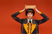 happy college girl in uniform holding stack of books on hand on orange background, knowledge puzzle #712420286