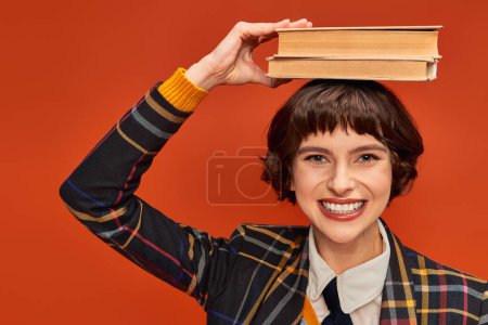 positive college girl in uniform holding stack of books on hand on orange background, knowledge