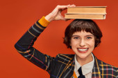 positive college girl in uniform holding stack of books on hand on orange background, knowledge hoodie #712420362