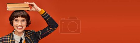 Photo for Positive college girl in uniform holding stack of books on hand on orange background, banner - Royalty Free Image