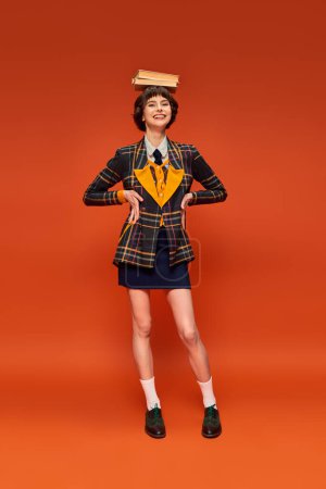 graceful and happy college girl in uniform holding stack of books on hand on orange background Poster 712420410