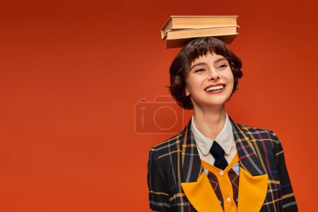 Foto de Optimista chica universitaria en uniforme celebración pila de libros en la mano sobre fondo naranja - Imagen libre de derechos