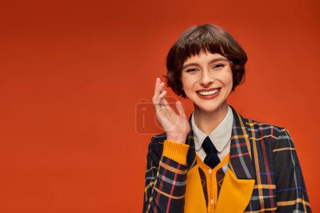 Photo for Optimistic college girl in checkered uniform waving hand on orange background, happy student life - Royalty Free Image