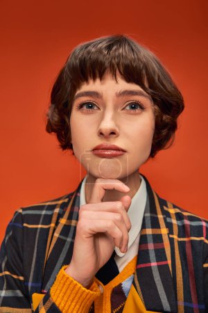 thoughtful college girl with short hair posing in checkered uniform on orange background