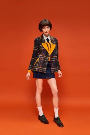 College-Mädchen in voller Länge in kariertem Blazer und Schuhen mit Socken auf orangefarbenem Hintergrund