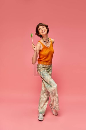 happy and funny young woman in vibrant attire holding her lollipop on pink background, candy lover