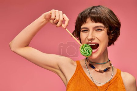 Photo for Positive young woman in orange knitted tank top biting colorful sweet lollipop on pink backdrop - Royalty Free Image