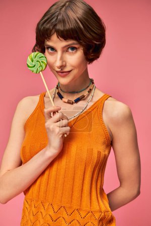brunette young woman in orange knitted tank top holding colorful sweet lollipop on pink backdrop