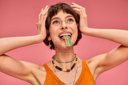 glückliche junge Frau schmeckt süß-saure Bonbonstreifen auf der Zunge, rosa Hintergrund
