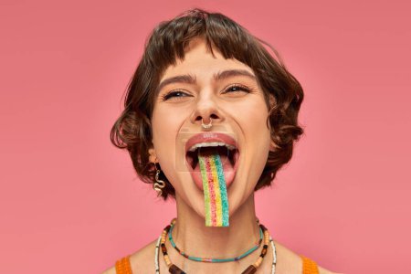 Téléchargez les photos : Joyeuse jeune femme dans la vingtaine dégustation sucrée et aigre bande de bonbons sur sa langue, fond rose - en image libre de droit