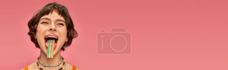 Téléchargez les photos : Bannière de jeune femme heureuse dégustation bande de bonbons aigre-douce sur sa langue, fond rose - en image libre de droit
