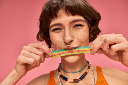 portrait of cheerful young woman in her 20s tasting sweet and sour candy strip on pink background
