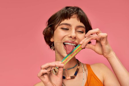 fröhliche junge Frau in ihren Zwanzigern leckt süß-saure Bonbonstreifen auf der Zunge, rosa Hintergrund