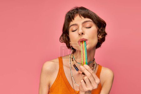 Porträt einer fröhlichen jungen Frau in ihren 20er Jahren, die süß-saure Bonbonstreifen auf rosa Hintergrund beißt