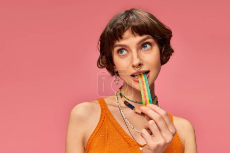 Téléchargez les photos : Portrait de jeune femme ludique dans sa vingtaine mordant bande de bonbons aigre-douce sur fond rose - en image libre de droit
