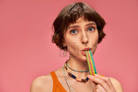 Porträt einer verspielten Frau in ihren 20er Jahren, die süß-saure Bonbonstreifen auf rosa Hintergrund beißt