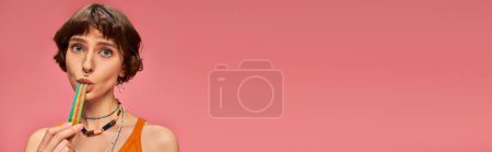portrait of playful woman in her 20s biting sweet and sour candy strip on pink background, banner