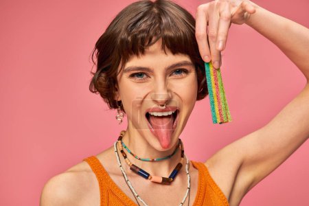 Téléchargez les photos : Femme heureuse dans sa vingtaine tenant sucré et aigre bande de bonbons et collant langue sur fond rose - en image libre de droit