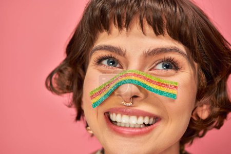 Téléchargez les photos : Femme heureuse dans sa vingtaine avec sucré et aigre bande de bonbons aon son nez sur fond rose - en image libre de droit