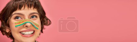 happy woman in her 20s with sweet and sour candy strip aon her nose on pink background, banner