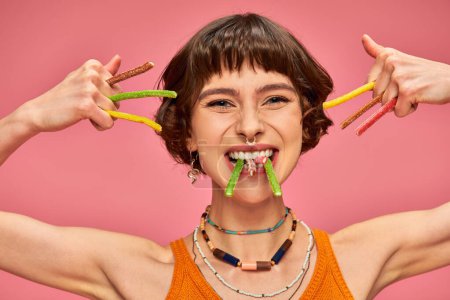 Photo for Happy young woman in her 20s with sweet and sour candies in hands and teeth on pink background - Royalty Free Image