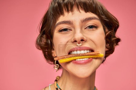 close up of happy woman in her 20s with sweet and sour candies in her white teeth on pink background