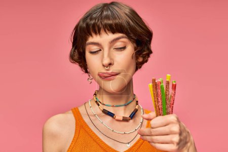 Téléchargez les photos : Ludique jeune femme dans sa vingtaine collant la langue et tenant bonbons aigre-doux à la main sur rose - en image libre de droit
