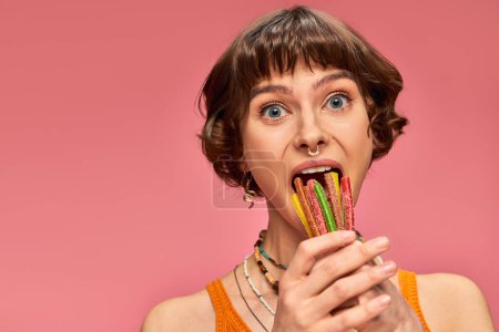 divertida joven de 20 años poniendo un montón de dulces y amargos en su boca abierta en rosa