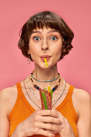 Photo for Cute pierced woman in her 20s smiling and holding bunch of sweet and sour candies in hands on pink - Royalty Free Image