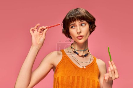 Téléchargez les photos : Douteuse fille percée dans sa vingtaine de manger deux saveurs différentes de bonbons aigre-doux sur rose - en image libre de droit