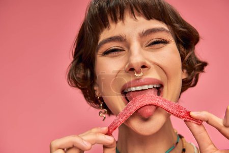 happy and young woman with nose piercing licking sweet and sour candy strip on pink background