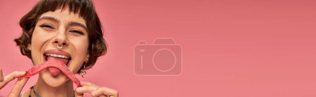 Téléchargez les photos : Heureuse jeune femme avec nez percing léchage sucré et aigre bande de bonbons sur fond rose, bannière - en image libre de droit