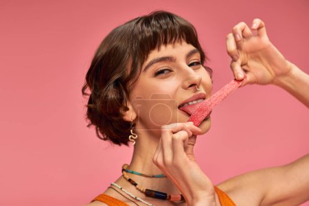fröhliche und junge Frau mit Nasenpiercing leckt süßen und sauren Bonbonstreifen auf rosa Hintergrund