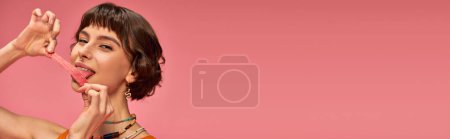Téléchargez les photos : Heureuse fille avec nez percing lécher sucré et aigre bande de bonbons sur fond rose, horizontal - en image libre de droit