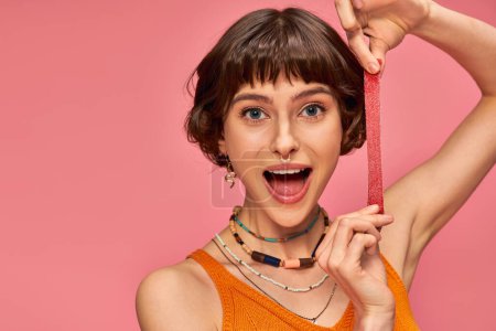 mujer excitada y joven con piercing en la nariz sosteniendo dulce y amarga tira de caramelo sobre fondo rosa