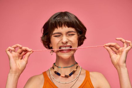 expressive and young woman with nose piercing biting sweet and sour candy strip on pink background