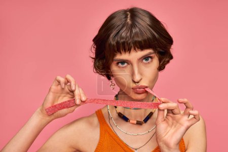 brünette und junge Frau mit Nasenpiercing beißenden süßen und sauren Bonbonstreifen auf rosa Hintergrund