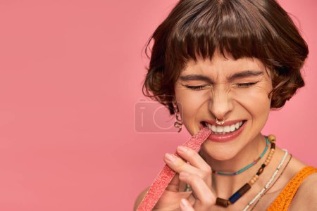 Photo for Expressive woman with nose and white teeth biting sweet and sour candy strip on pink - Royalty Free Image