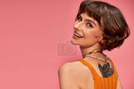 happy tattooed woman in 20s with nose piercing and brunette short hair smiling on pink background