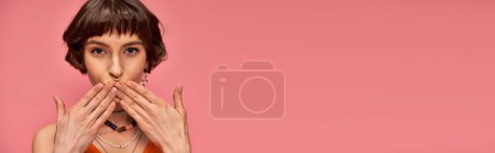 pretty woman with nose piercing and short brunette hair sending air kiss on pink background, banner