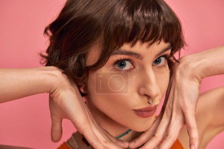 flirty young woman with nose piercing and short brunette hair pouting lips on pink background