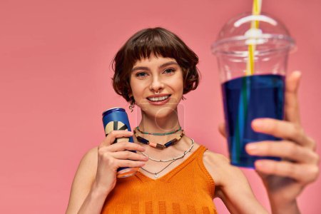 Foto de Mujer joven positiva con el pelo corto morena y piercing celebración de bebidas de verano en el telón de fondo rosa - Imagen libre de derechos