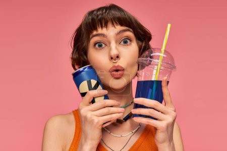 Photo for Amazed young woman with short brunette hair and piercing holding summer drinks on pink backdrop - Royalty Free Image