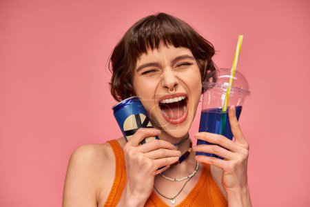 Foto de Mujer joven excitada con el pelo corto morena y piercing celebración de bebidas de verano sobre fondo rosa - Imagen libre de derechos