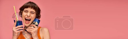 Photo for Excited young woman with short brunette hair and piercing holding summer drinks on pink, banner - Royalty Free Image
