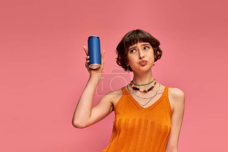 jeune femme incertaine aux cheveux bruns courts et piercing tenant une canette de soda sur rose, boisson d'été