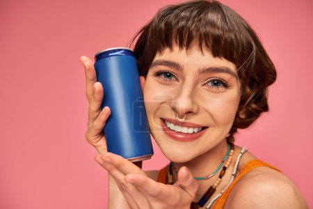 close up of joyful young woman with short brunette hair holding soda can on pink, summer beverage