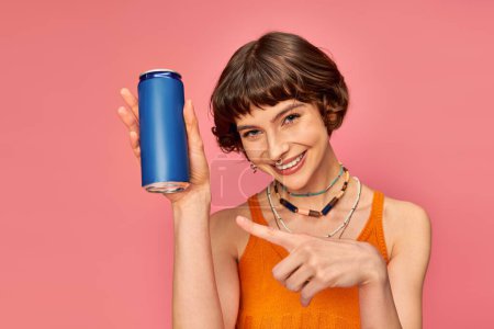 Téléchargez les photos : Joyeuse jeune femme aux cheveux bruns courts pointant vers la canette de soda sur rose, boisson d'été - en image libre de droit