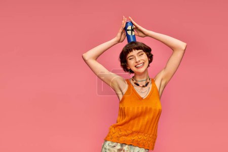 Téléchargez les photos : Joyeuse jeune femme aux cheveux bruns courts posant avec de la soude peut sur sa tête sur rose, boisson d'été - en image libre de droit