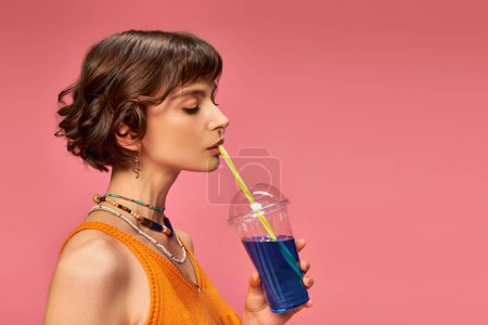 Téléchargez les photos : Jeune femme aux cheveux bruns courts boire cocktail d'été rafraîchissant de paille sur rose - en image libre de droit